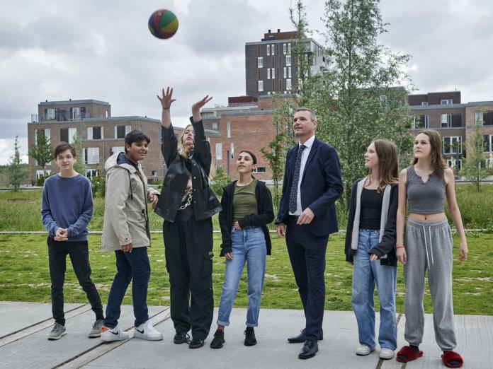 Jakob Næsager står i en skolegård med en gruppe store børn. En pige spiller volleyball.